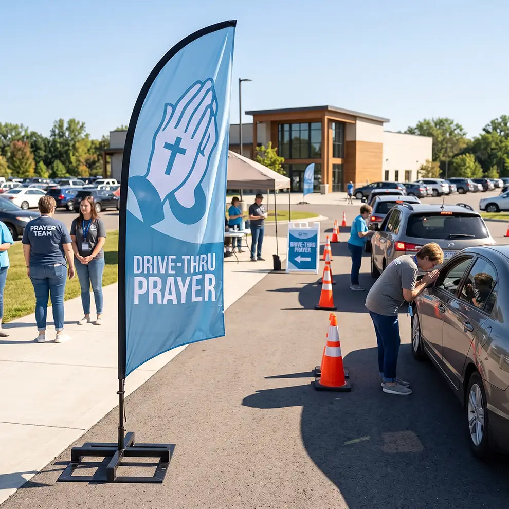 A tall blue feather flag with a praying hands graphic and the words 'DRIVE-THRU PRAYER' stands on a sidewalk beside a drive-thru lane at a church or community center. People are standing and checking cars, with orange traffic cones guiding the flow.