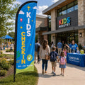 Kids ministry check-in church feather flag outside a busy family ministry entrance with parents, children, and volunteers