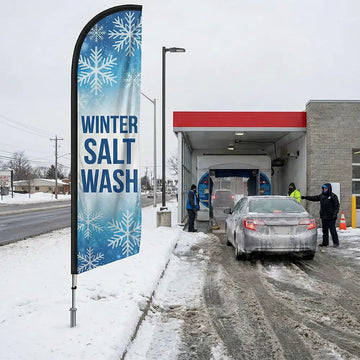 Winter Salt Wash feather flag with blue and white snowflake graphics positioned on a snowy roadside.